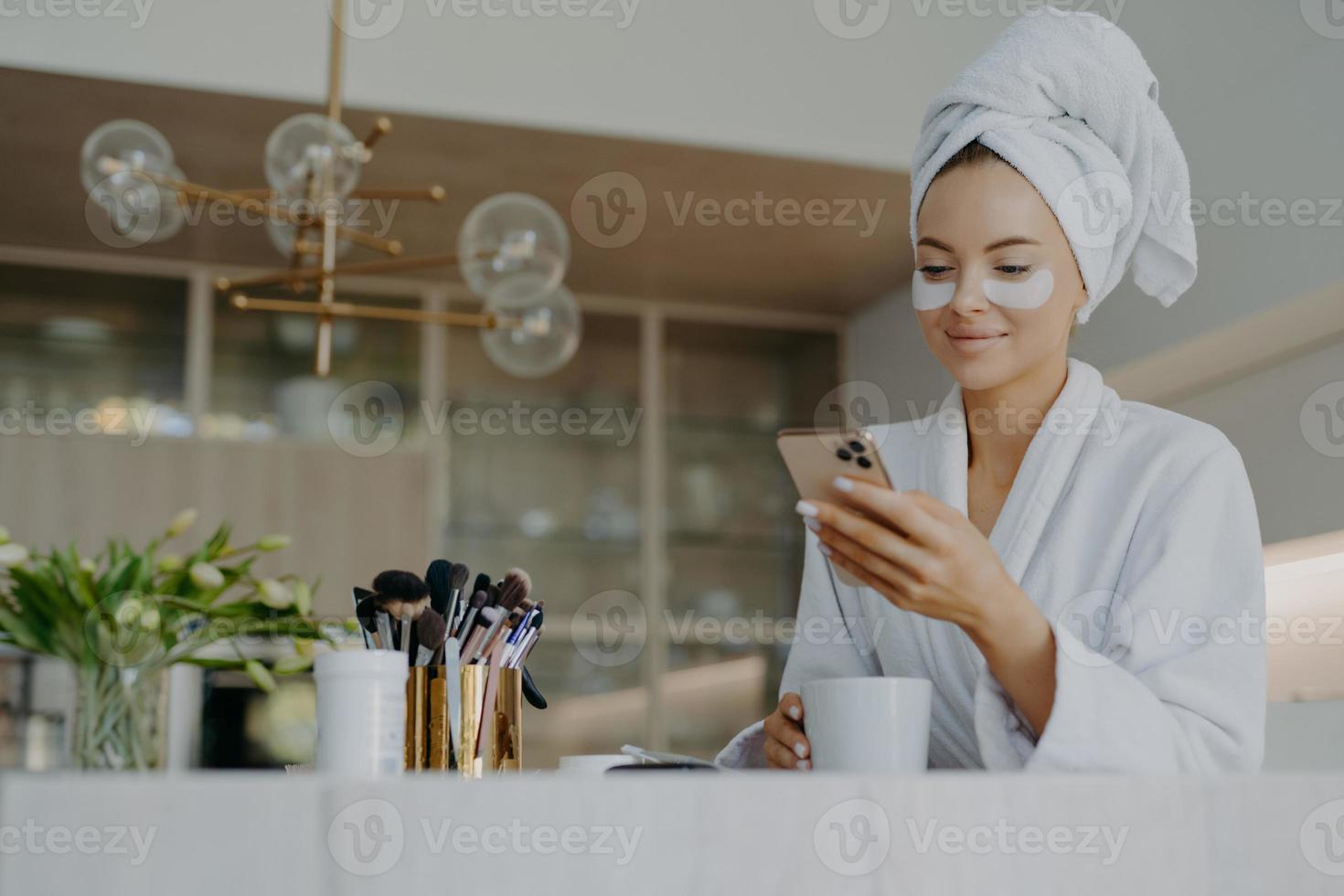 Photo of attractive young woman wears bath towel on head and soft white dressing gown holds mobile phone enjoys online communication applies moisturising undereye patches drinks hot beverage