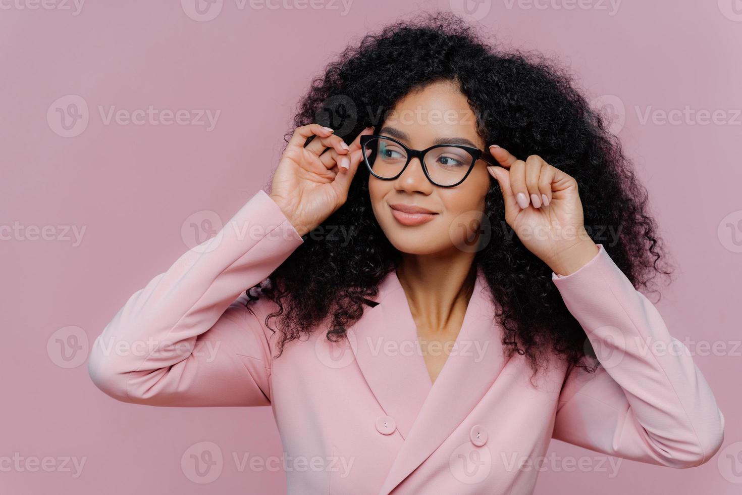 Headshot of attentive female boss keeps hands on frame of glasses, looks pensively away, wears rosy formal suit, has curly Afro hairstyle, poses against purple background. People, business, ethnicty photo