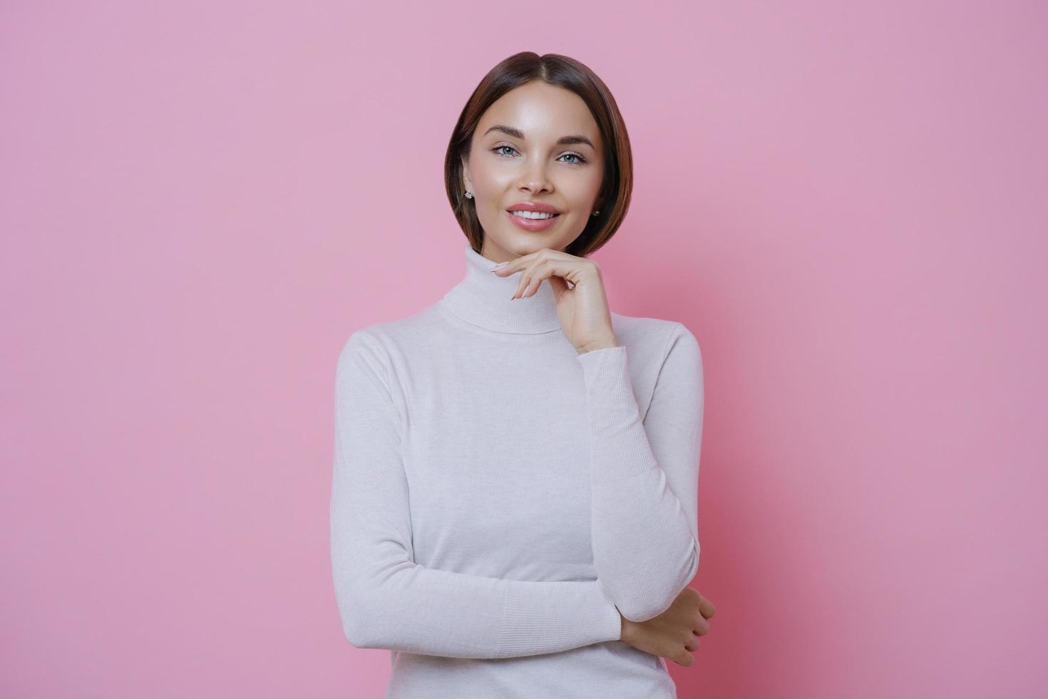 Young beautiful woman with pleased expression, touches chin, smiles tenderly, has straight dark hair, dressed in casual white turtleneck, isolated on pink background. Femininity, beauty and skin care photo