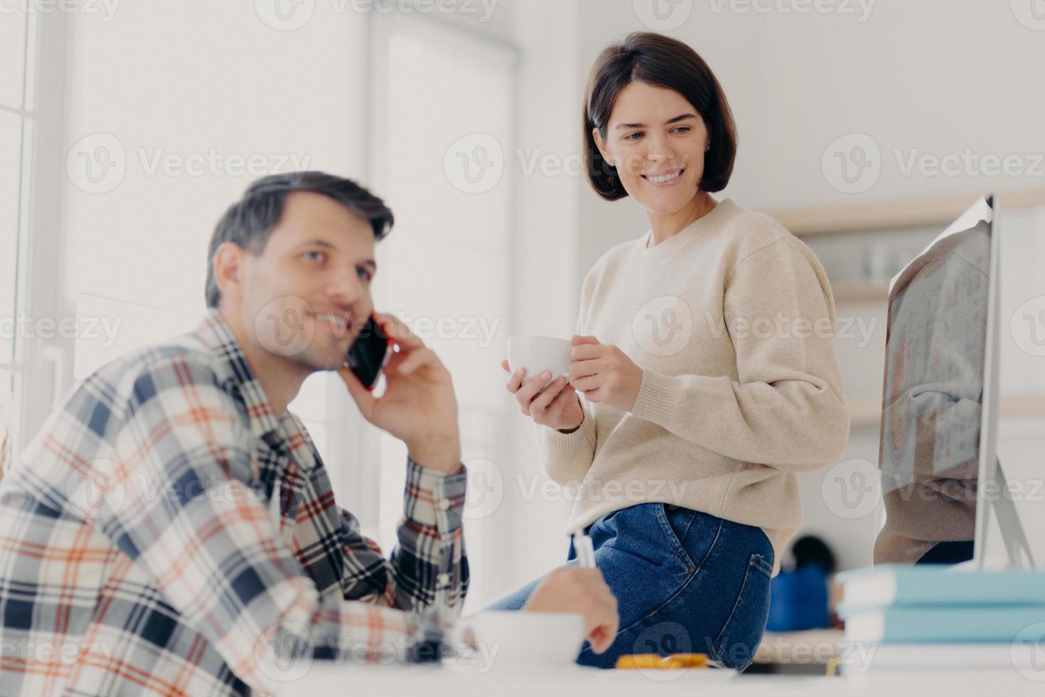 Young married couple study contract for new house, check documents for buying property, man discusses something with partner via cellphone, happy woman drinks tea and looks in computer monitor photo