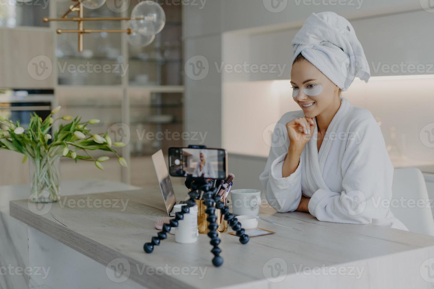 Beautiful successful female blogger records tutorial video talks with followers looks at camera of smartphone on tripod applies beauty patches under eyes dicuss how to care about skin and complexion photo