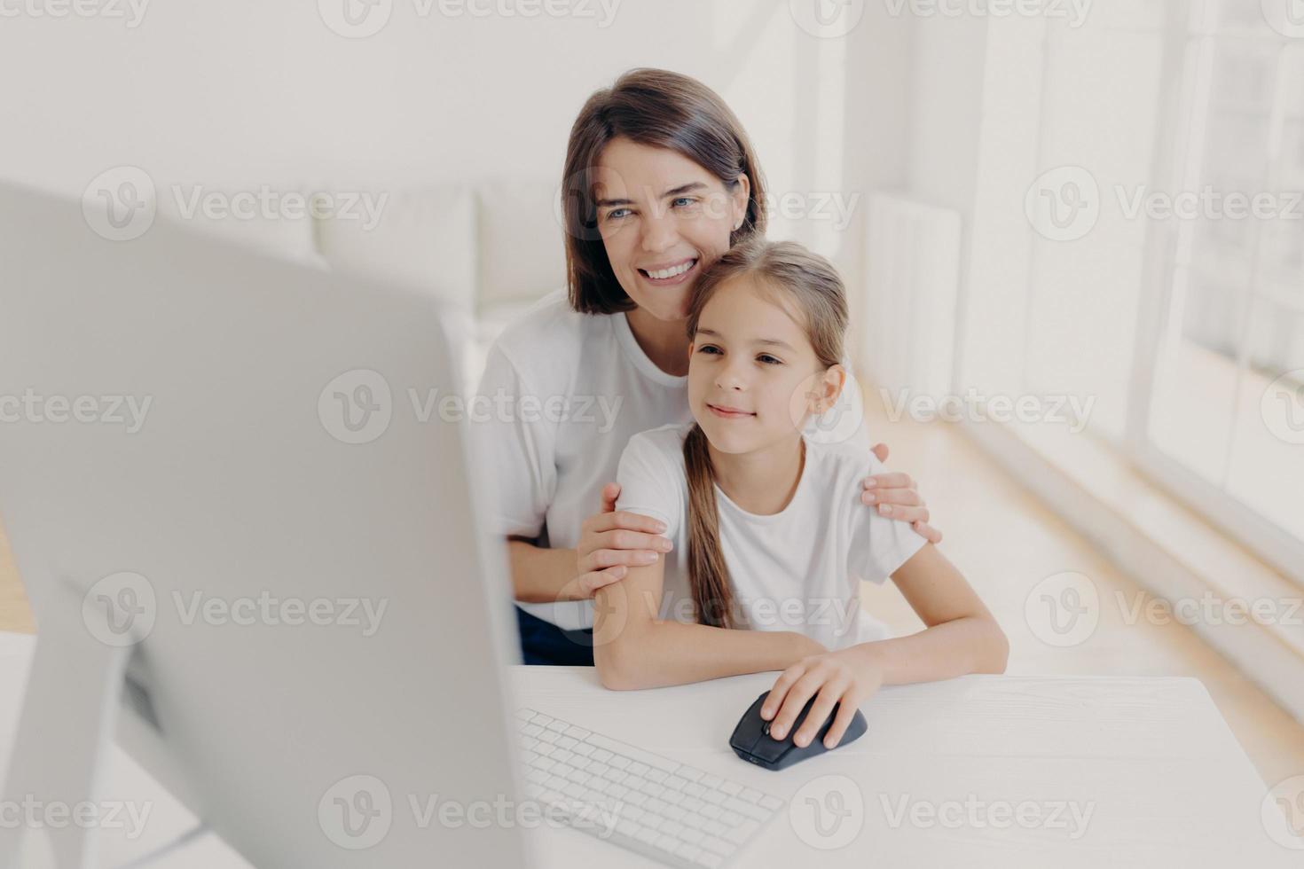 Happy affectionate mom embraces her small daughter, work together on modern computer, preapare presentation for lesson, look gladfully into monitor, spend leisure time at home, surf internet page. photo