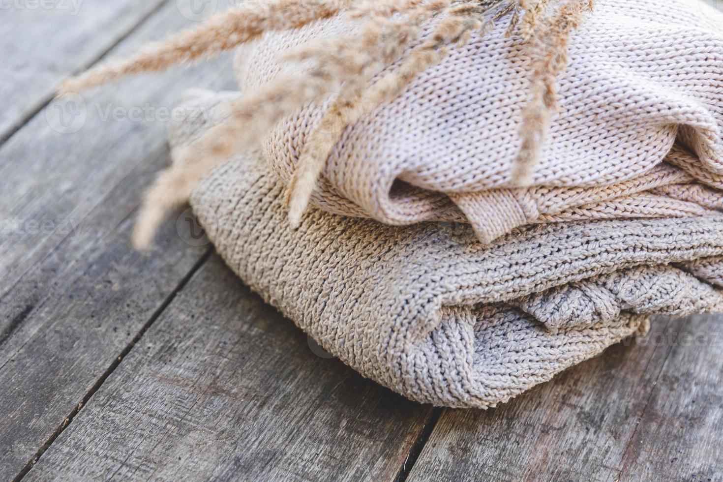 Autumn or winter background. Stack of cozy knitted wool clothes sweaters and dried plant on wooden table. Hygge mood, cold weather concept. Handmade knitted sweaters. photo