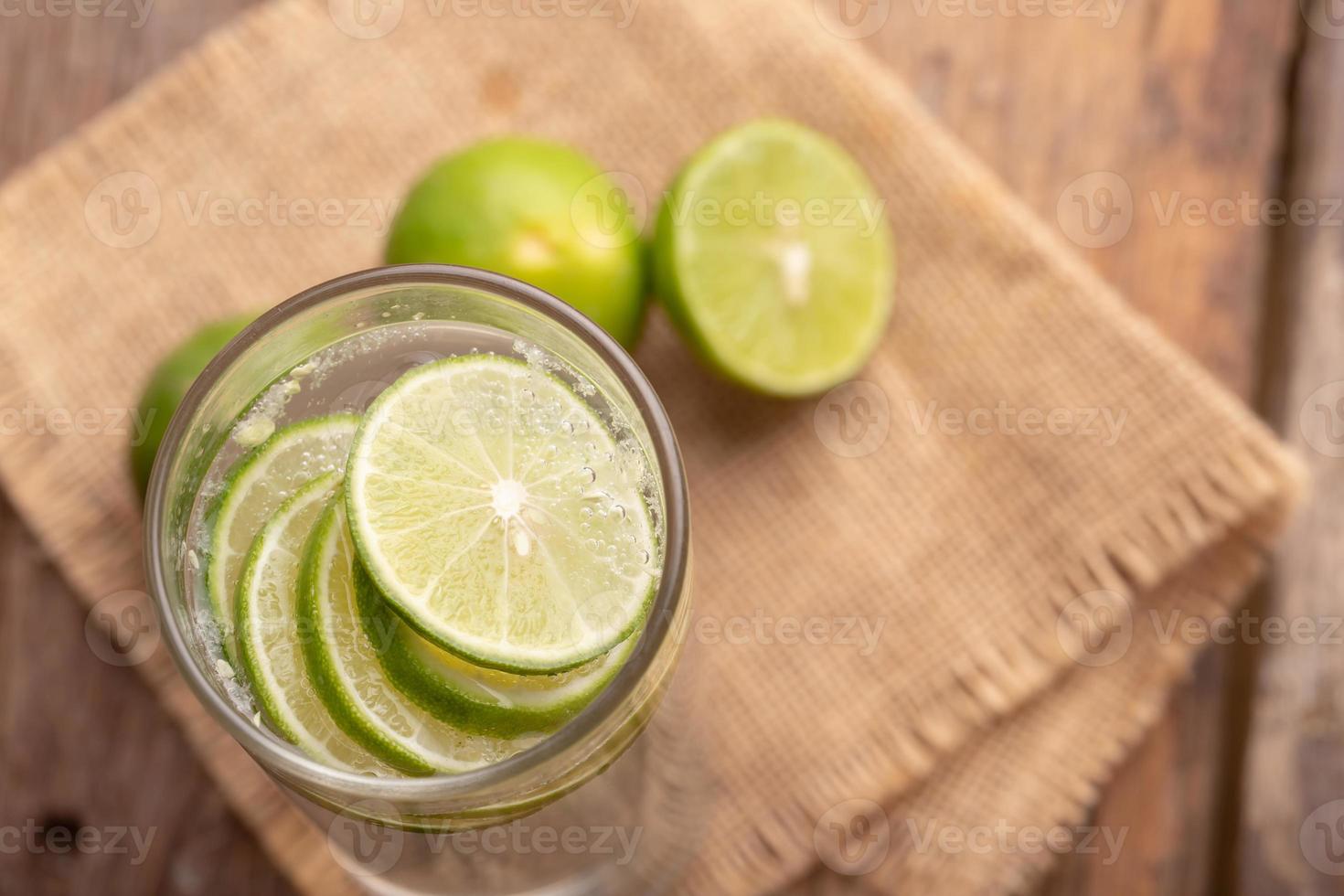 cierre el limón cortado en rodajas en el vaso con agua de soda y la mitad del lugar de lima verde en un saco tejido y una mesa de madera foto