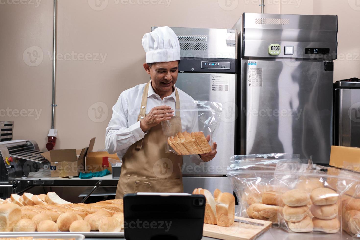 chef asiático senior en delantal, blogger de pequeñas empresas que transmite en vivo a través de tecnología de aplicaciones móviles, promoción de ventas de panadería en línea, pan fresco, pastelería y cocina comercial en la cocina. foto