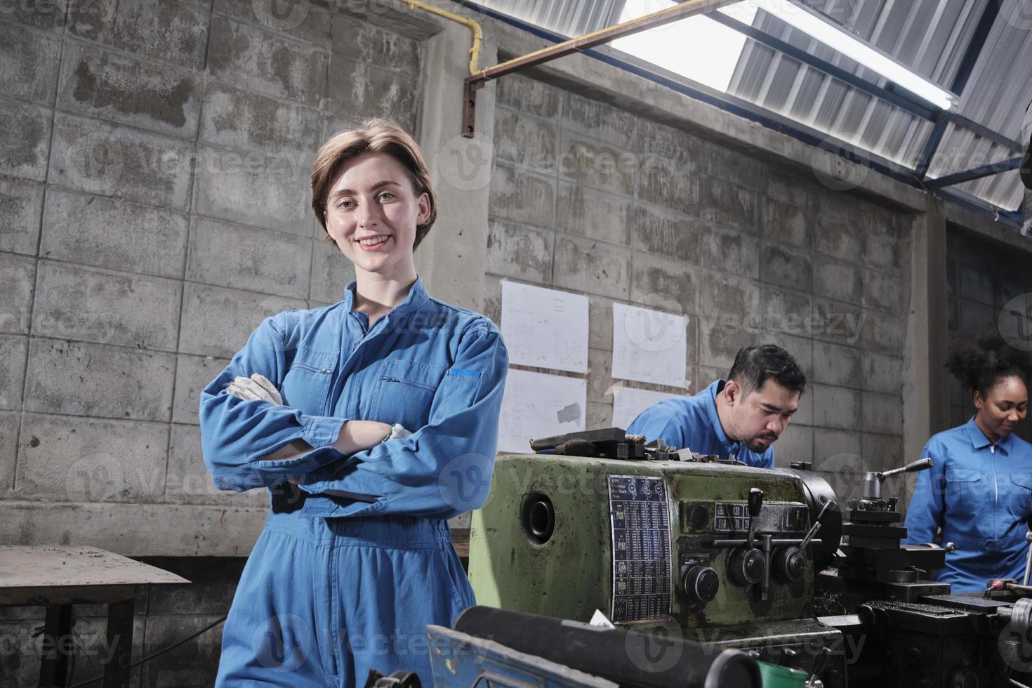 Portrait of Young White female industry worker stands, arms crossed, looks at camera and smiles, multiracial team works with machine behind in manufacturing factory, professional mechanical engineers. photo