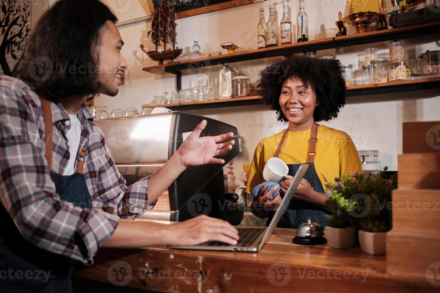 Two cafe business startup partners and friends, African American female, and Thai male baristas talk and cheerful smile together at counter bar of coffee shop, happy service job, and SME entrepreneur. photo