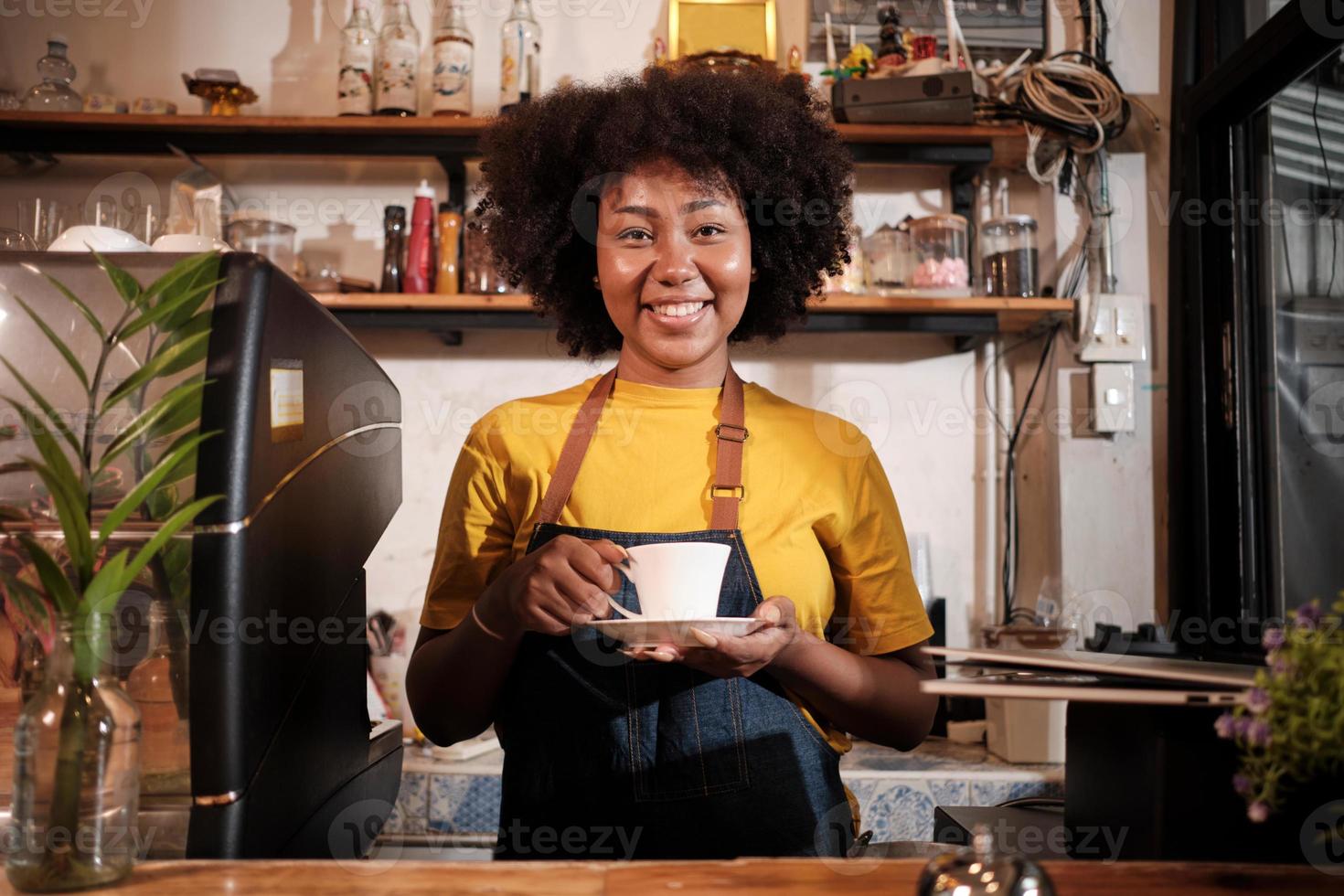 African American female barista in looks at camera, offers cup of coffee to customer with cheerful smile, happy service works in casual restaurant cafe, young small business startup entrepreneur. photo