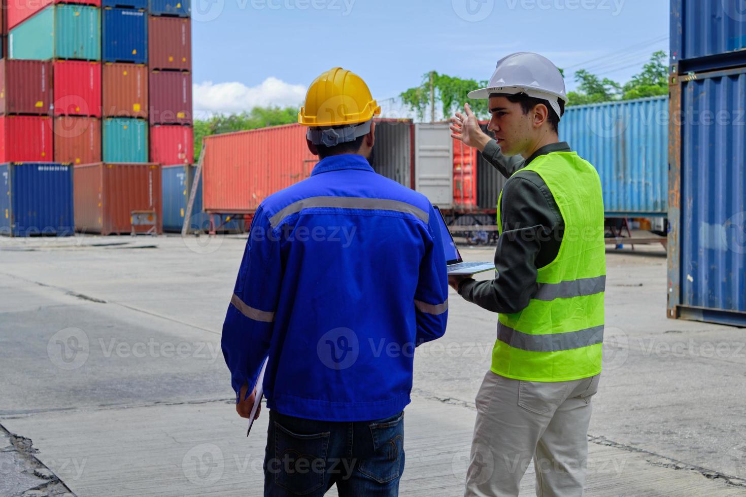 Two professional male workers in safety uniforms and hard hats work and inspect a logistics terminal with many stacks of containers, loading control shipping goods for cargo transportation industry. photo