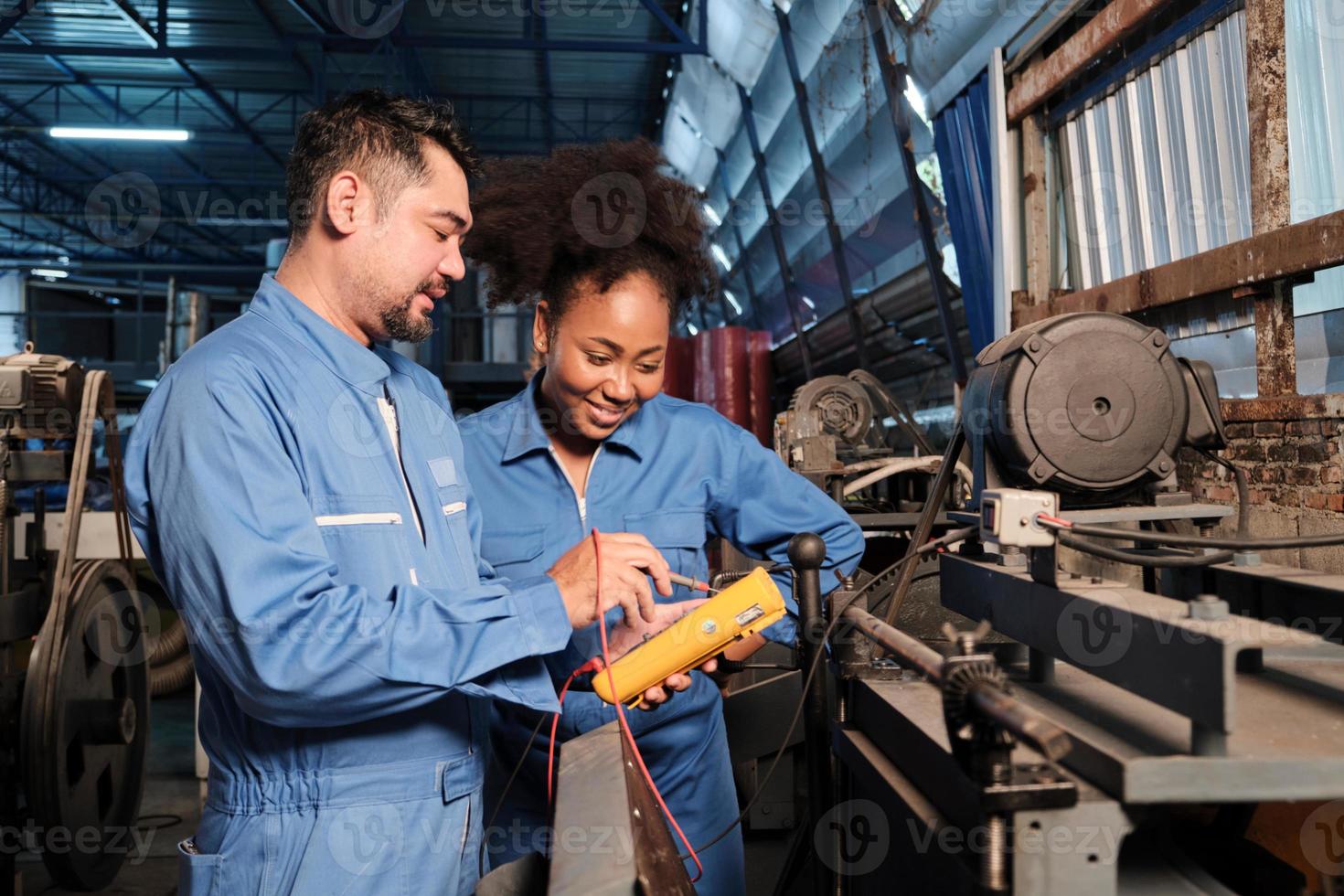 Asian male and female African American engineers in safety uniform work by inspecting machines' voltage current, checking, and maintaining at manufacture factory, electric system service occupations. photo