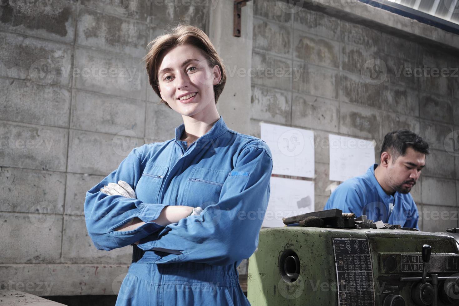 Portrait of Young White female industry worker stands, arms crossed, looks at camera and smiles, male partner works with machine behind her in manufacturing factory, professional mechanical engineers. photo