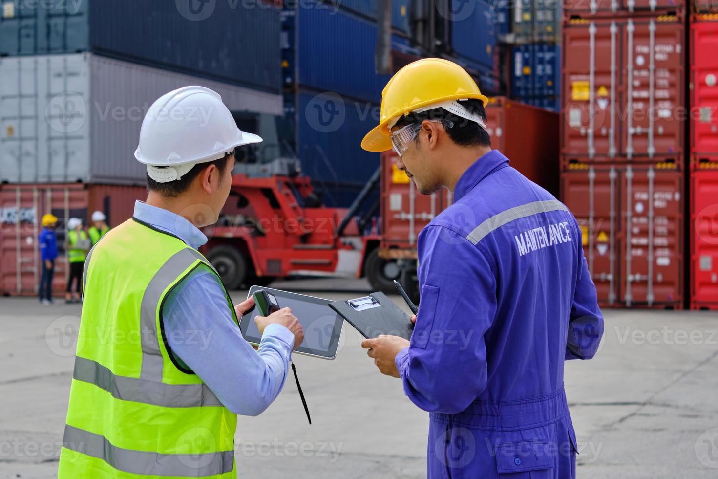 Two professional Asian male workers in safety uniforms and hard hats work at a logistics terminal with many stacks of containers, loading control shipping goods for the cargo transportation industry. photo