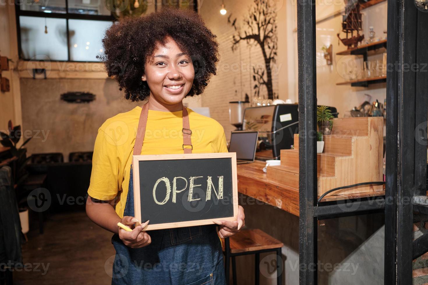 una barista nueva afroamericana se para en la puerta de un café informal, mira la cámara y muestra un cartel abierto, sonrisas felices y alegres con trabajos de servicio de cafetería y nuevos emprendedores de negocios. foto