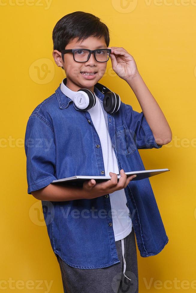 Close-up of smart little boy in casual clothes, headphones and eyeglasses holding book and smiling to the camera on yellow  background. photo