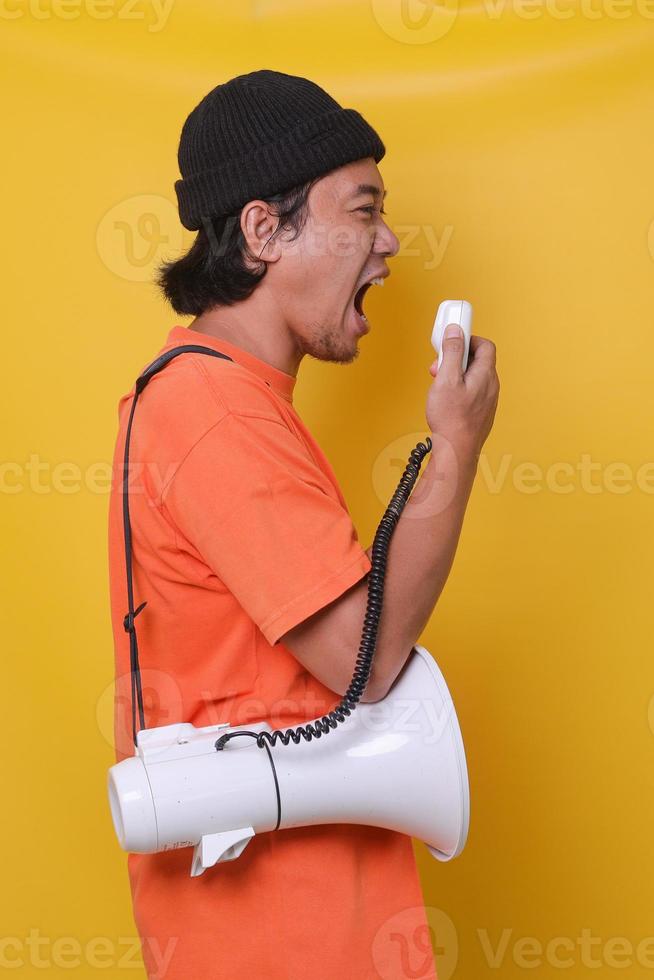 Close-up of Asian young man in casual style isolated on yellow background shouting through a megaphone. Side view. photo