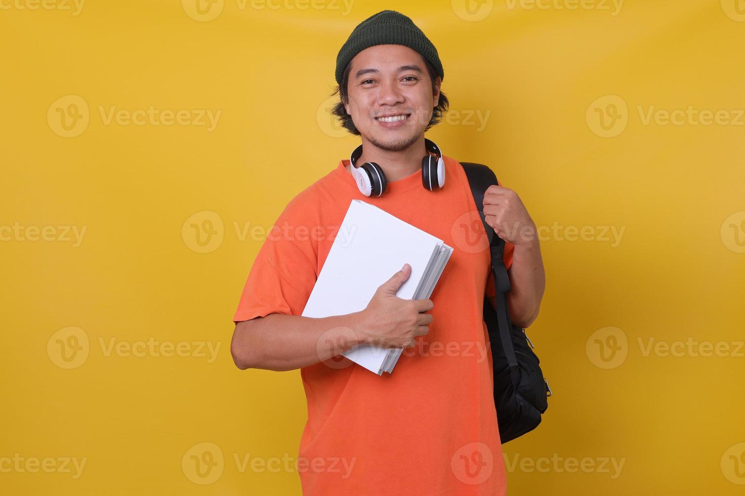 joven asiático de estilo informal con mochila y auriculares que traen libros aislados de fondo amarillo. concepto de escuela universitaria universitaria. foto