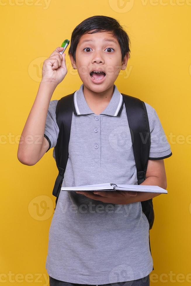 colegial asiático con una expresión cuando tiene una idea o una solución, vistiendo un polo gris y sosteniendo un libro y un bolígrafo. concepto de regreso a la escuela. foto