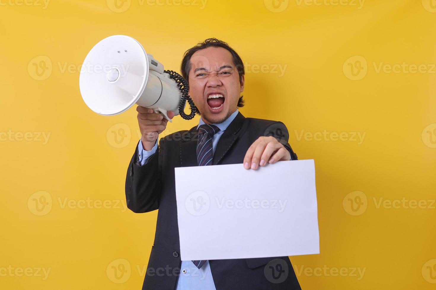 un hombre de negocios asiático está gritando y gritando usando un megáfono y sosteniendo una hoja de papel en blanco aislada en un fondo amarillo. copia espacio y maqueta. foto