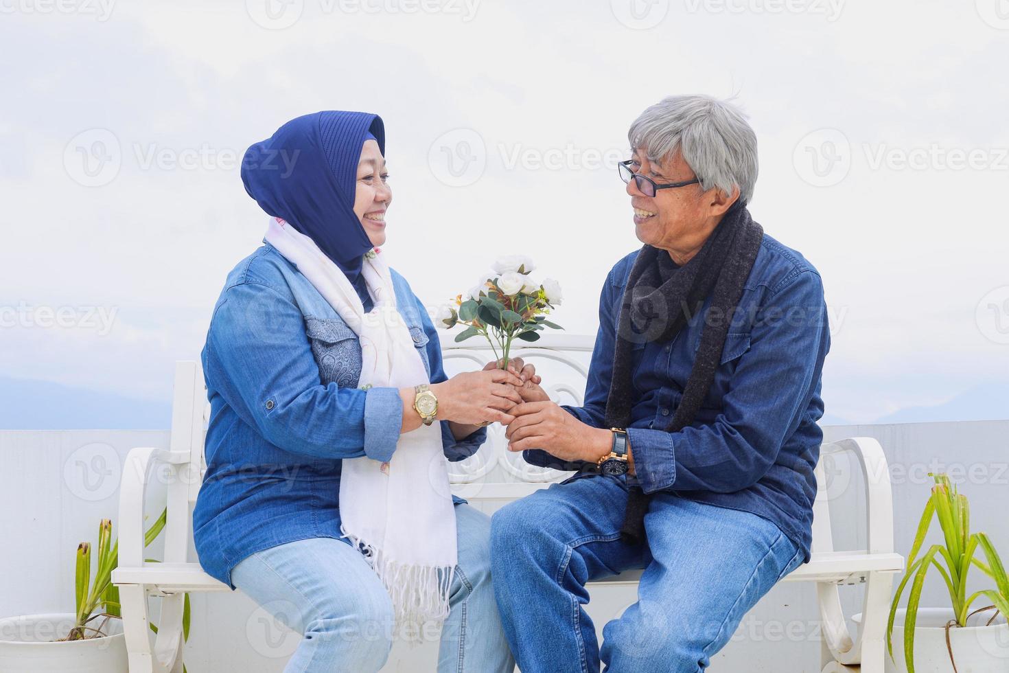 Asian Elderly couple. Joyful nice senior romantic couple is sitting and laughing each othet while holds together artificial white flower. photo