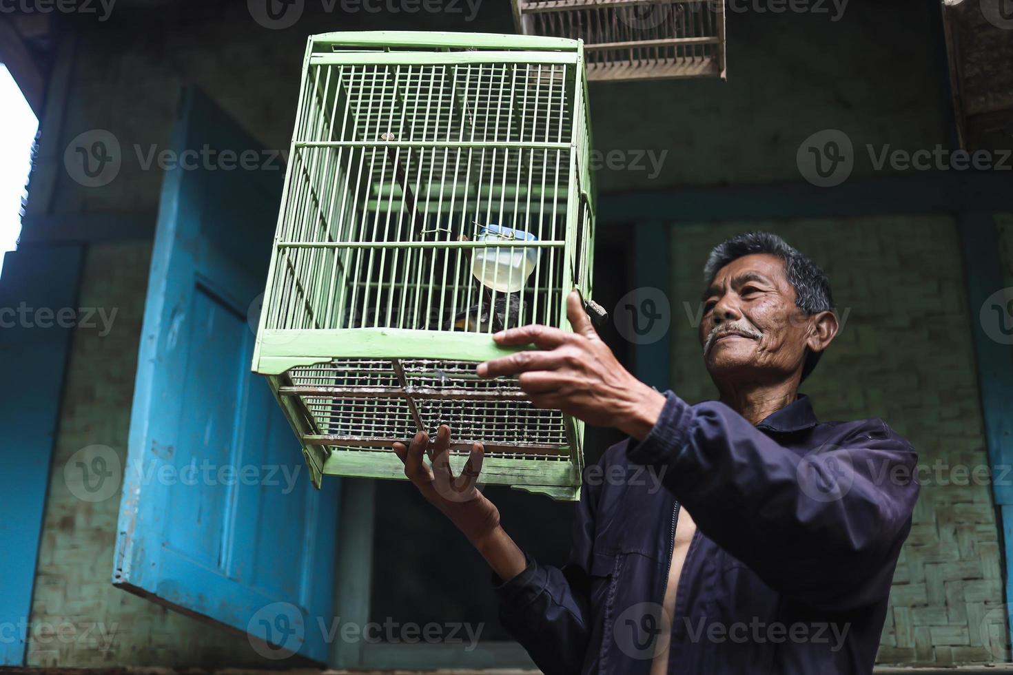 Asian elderly man holding green bird cage outside traditional wooden house. photo