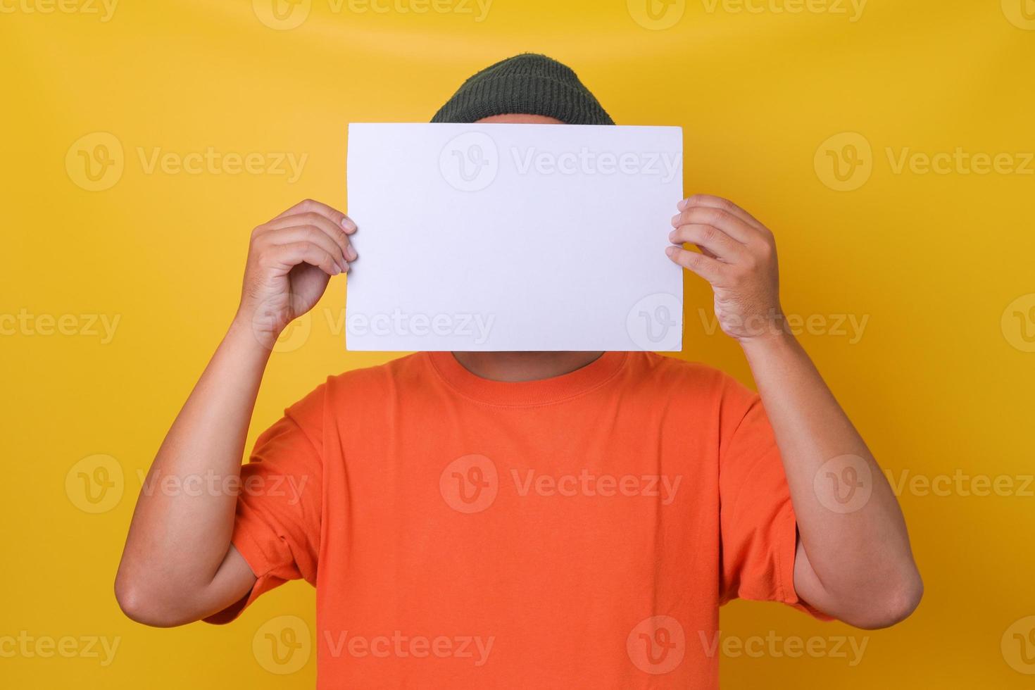 Young guy in orange t-shirt  hiding his face under lblank white paper, isolated yellow background. Horizontal, landscape style paper. photo