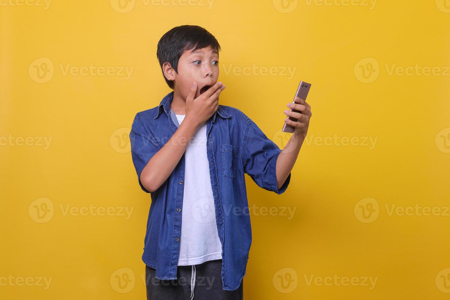 Asian boy in casual denim style is holding phone and showing surprised expression isolated on yellow background photo