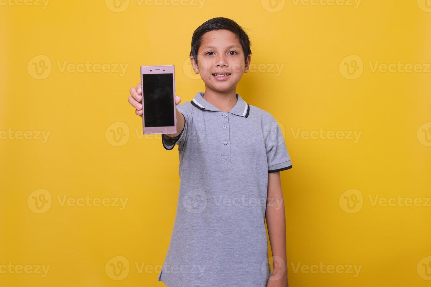 Asian boy in casual style is smiling and showing screen of smartphone isolated on yellow background. Copy space and mock up for online learning and kids fashion concept. photo