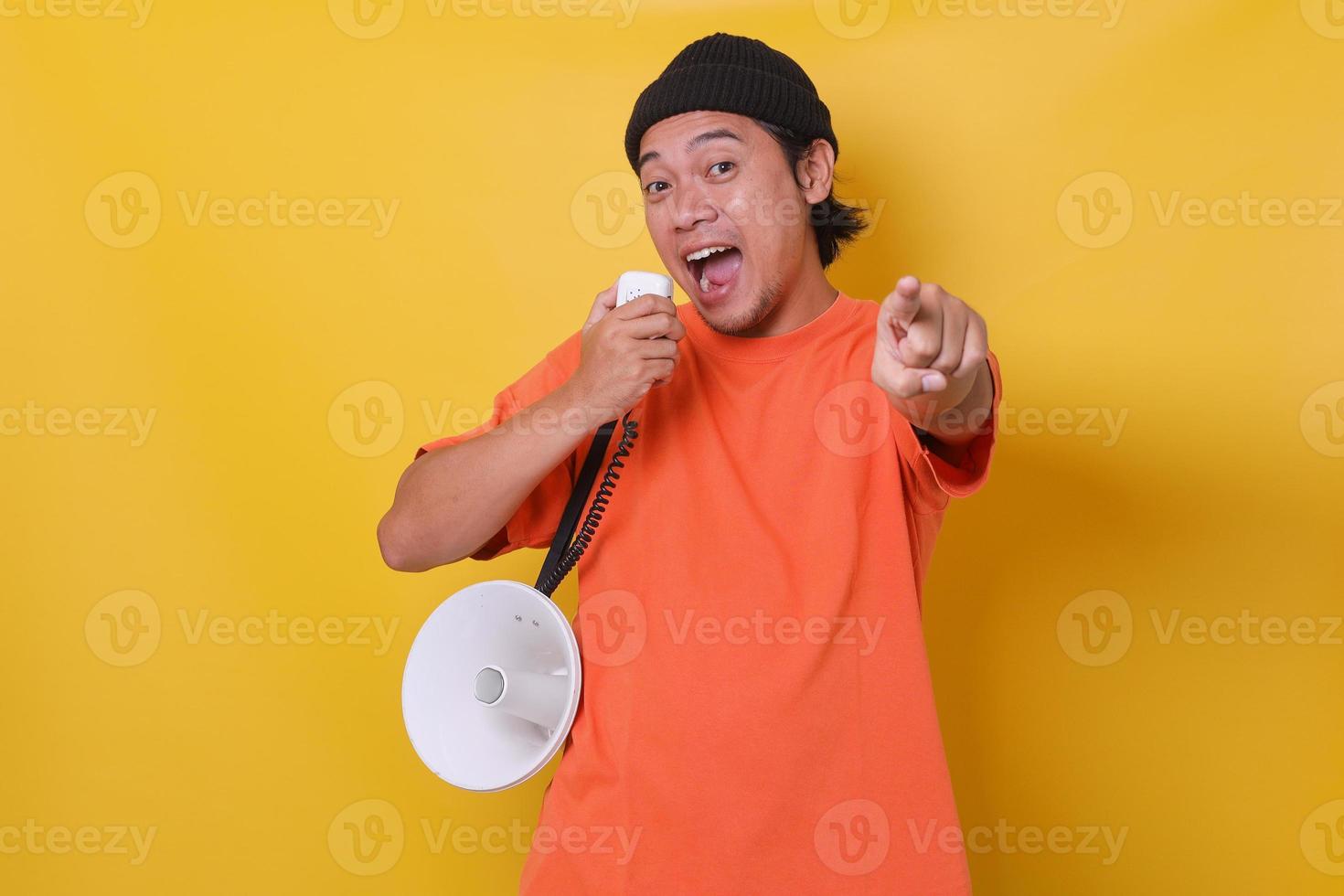 Asian young man in casual style using megaphone and pointing finger forward to the camera isolated on yellow background. photo