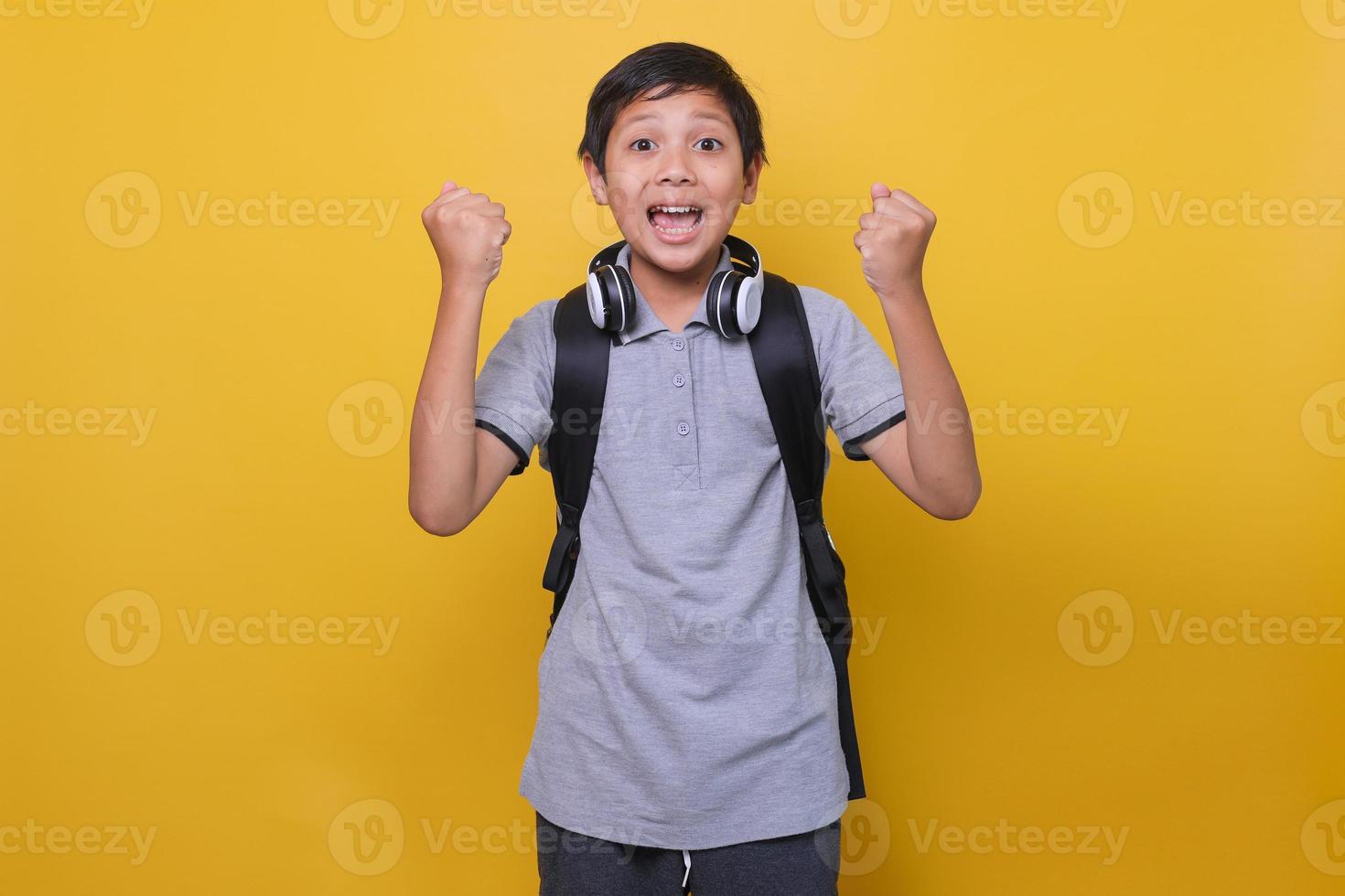Yes. Happy Asian school boy in casual style use black backpack and headphone shaking fists, making winner gesture, against yellow background. Back to school concept. photo
