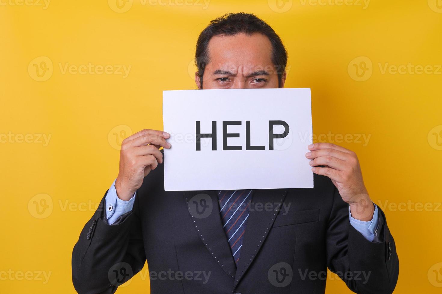 Asian man is frowning wearing business suit covering half his face with a paper written help, isolated on yellow background photo