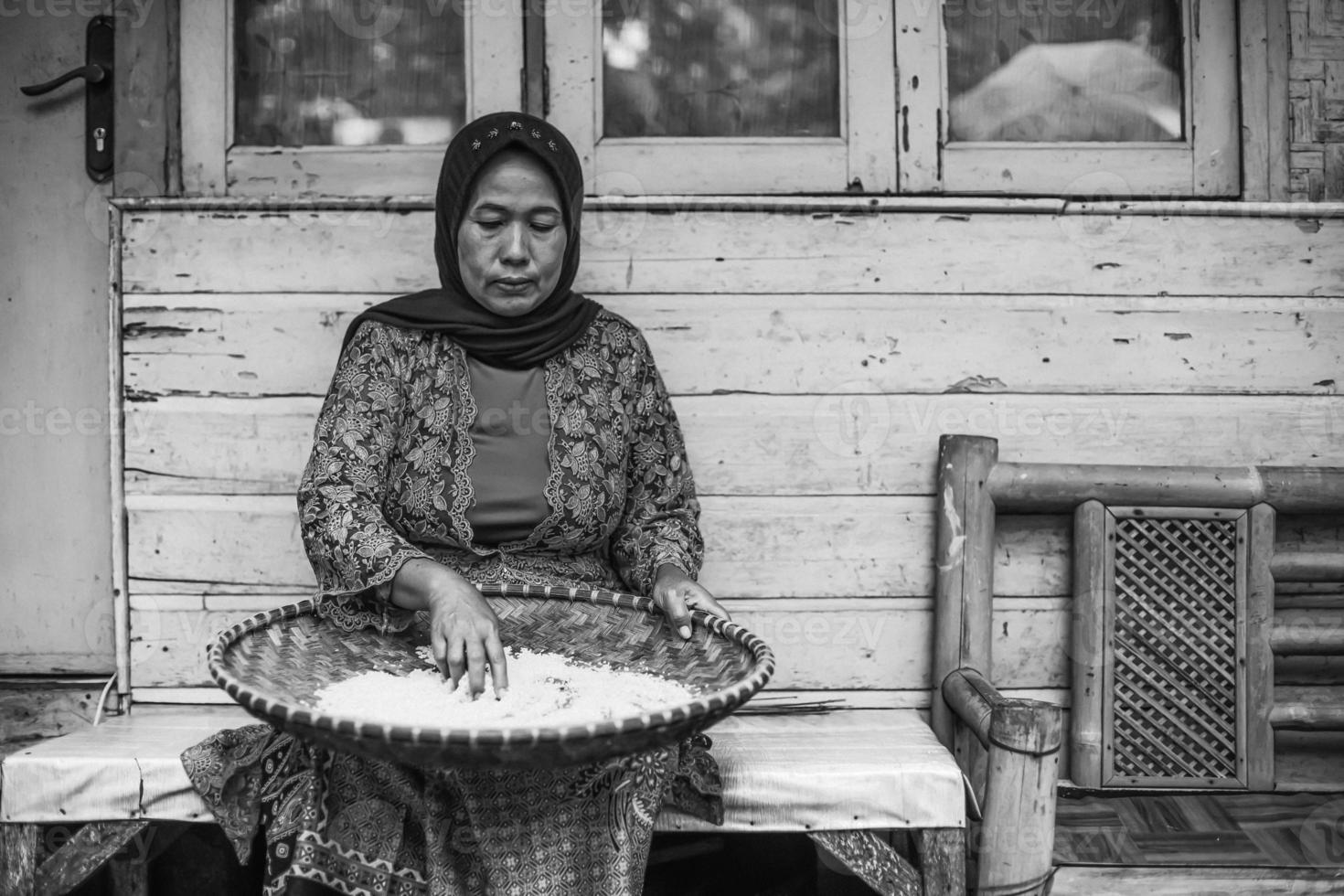 Black and white potrait of Asian local woman is sitting and sifting rice with bamboo colander against traditional wooden house. photo