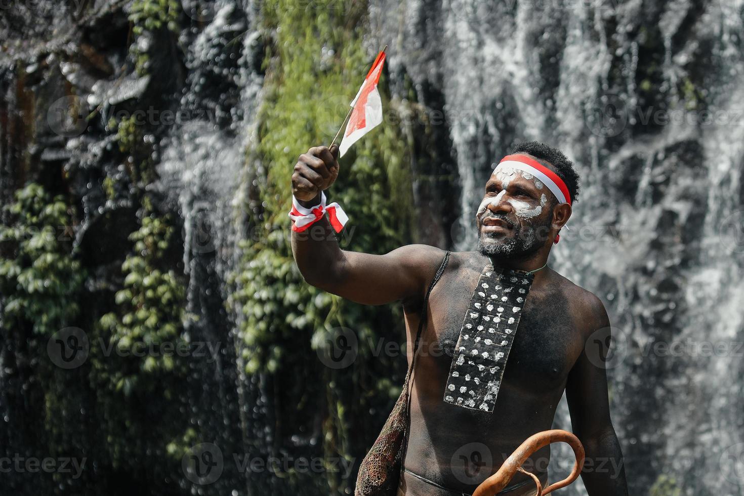 Spirit of Papua man wearing traditional clothes of Dani tribe, red-white headband and bangle is holding little Indonesia flag and celebrating Indonesia independence day against waterfall background. photo