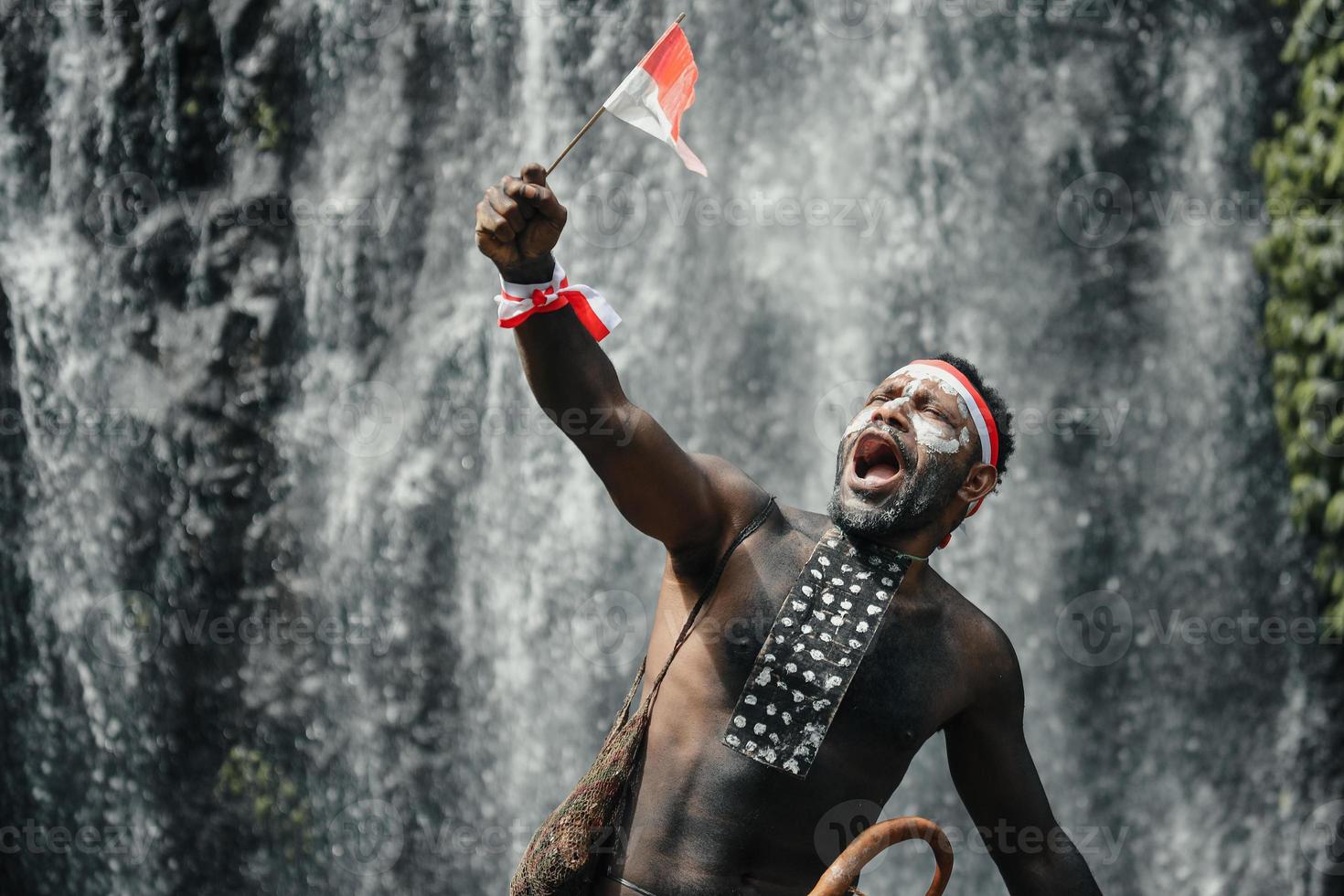 Papua man of Dani tribe say merdeka, celebrating Indonesia independence day against waterfall background. photo