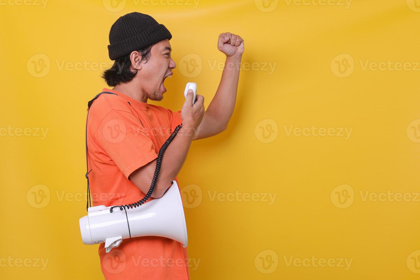 Asian young man in casual style giving spirit using megaphone isolated on yellow background. Side view. photo