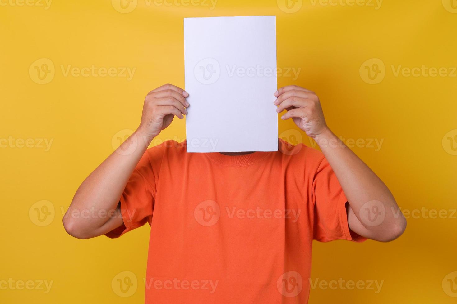 Young guy in orange t-shirt  hiding his face under blank white paper, isolated yellow background. photo