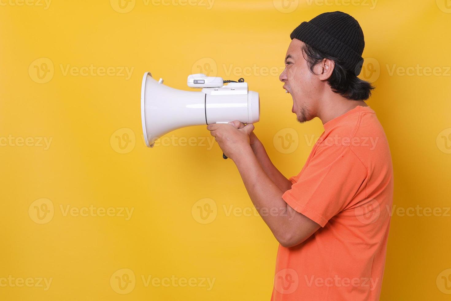 Asian young man in casual style isolated on yellow background shouting through a megaphone. Side view. photo