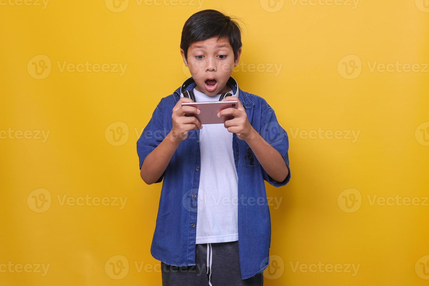 Cute Asian boy in casual denim style and headphones looking at his smartphone and feeling surprised and shocked. Studio shot on yellow background. photo