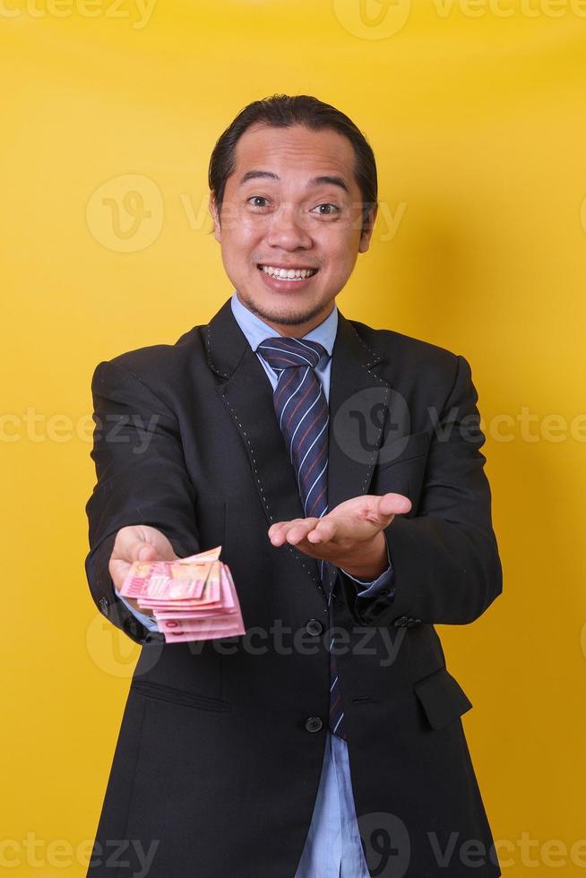 Attractive Asian man in business suit standing against yellow background, presenting and showing money one hundred thousand rupiah. Financial and savings concept. photo