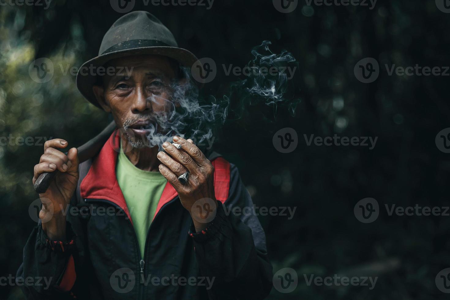 Close up of Asian farmer elderly man is smoking while carrying hoe after work on field photo