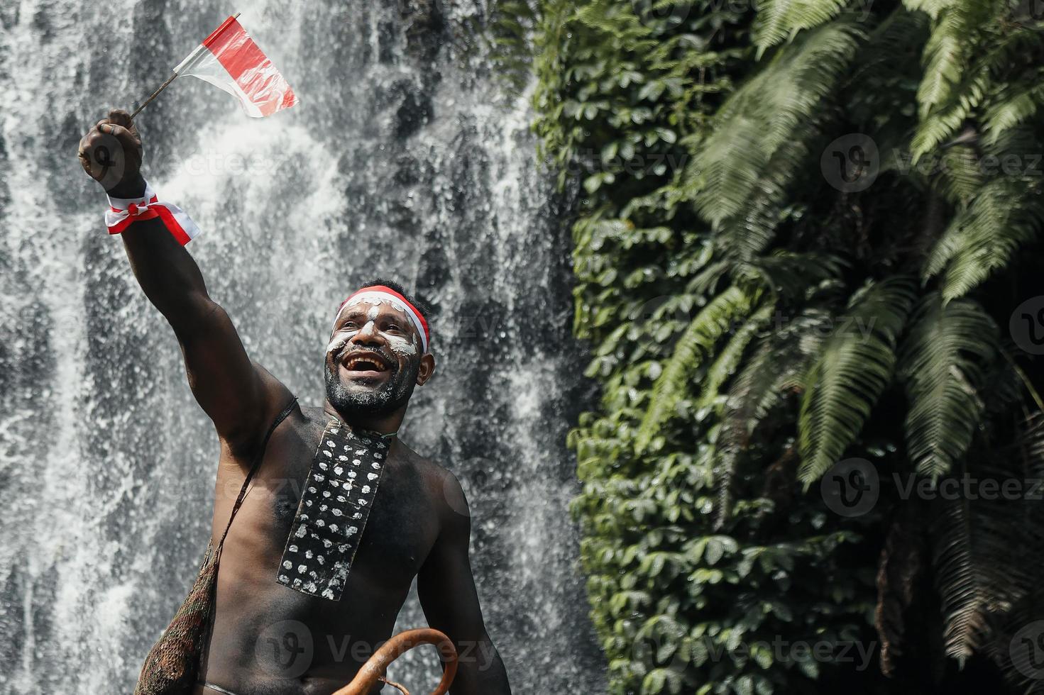 Spirit of Papua man wearing traditional clothes of Dani tribe, red-white headband and bangle is holding little Indonesia flag and celebrating Indonesia independence day against waterfall background. photo