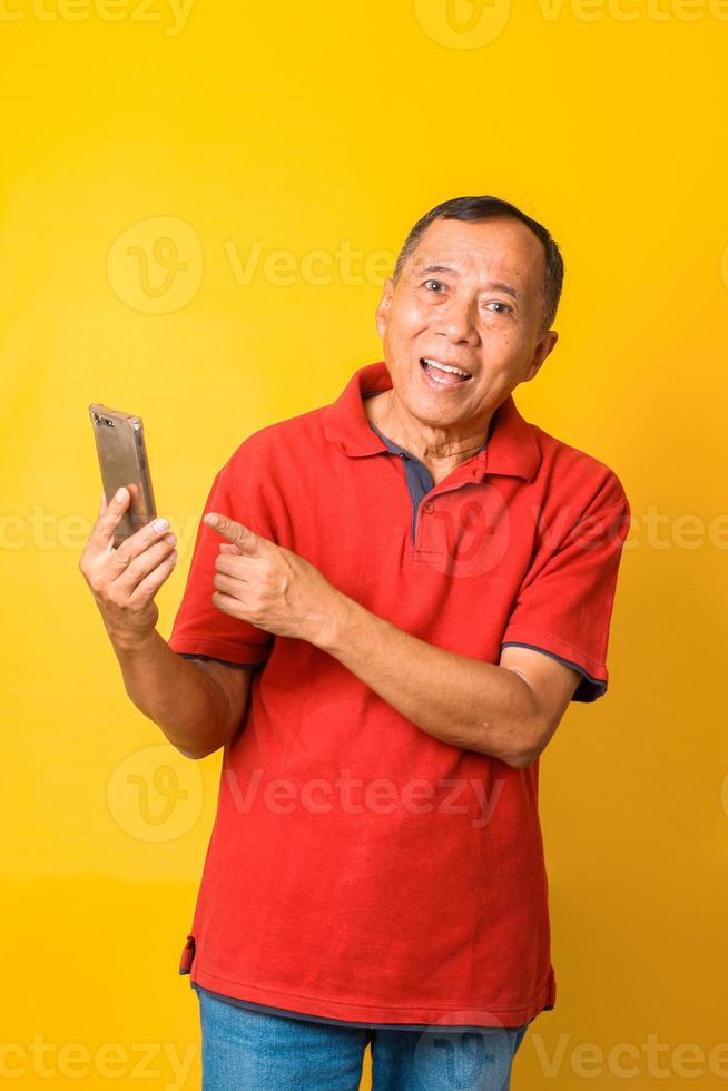 Photo of Asian old man smiling with wide eyes and holding smartphone with direct finger pointing to screen wearing red t-shirt isolated on yellow color background