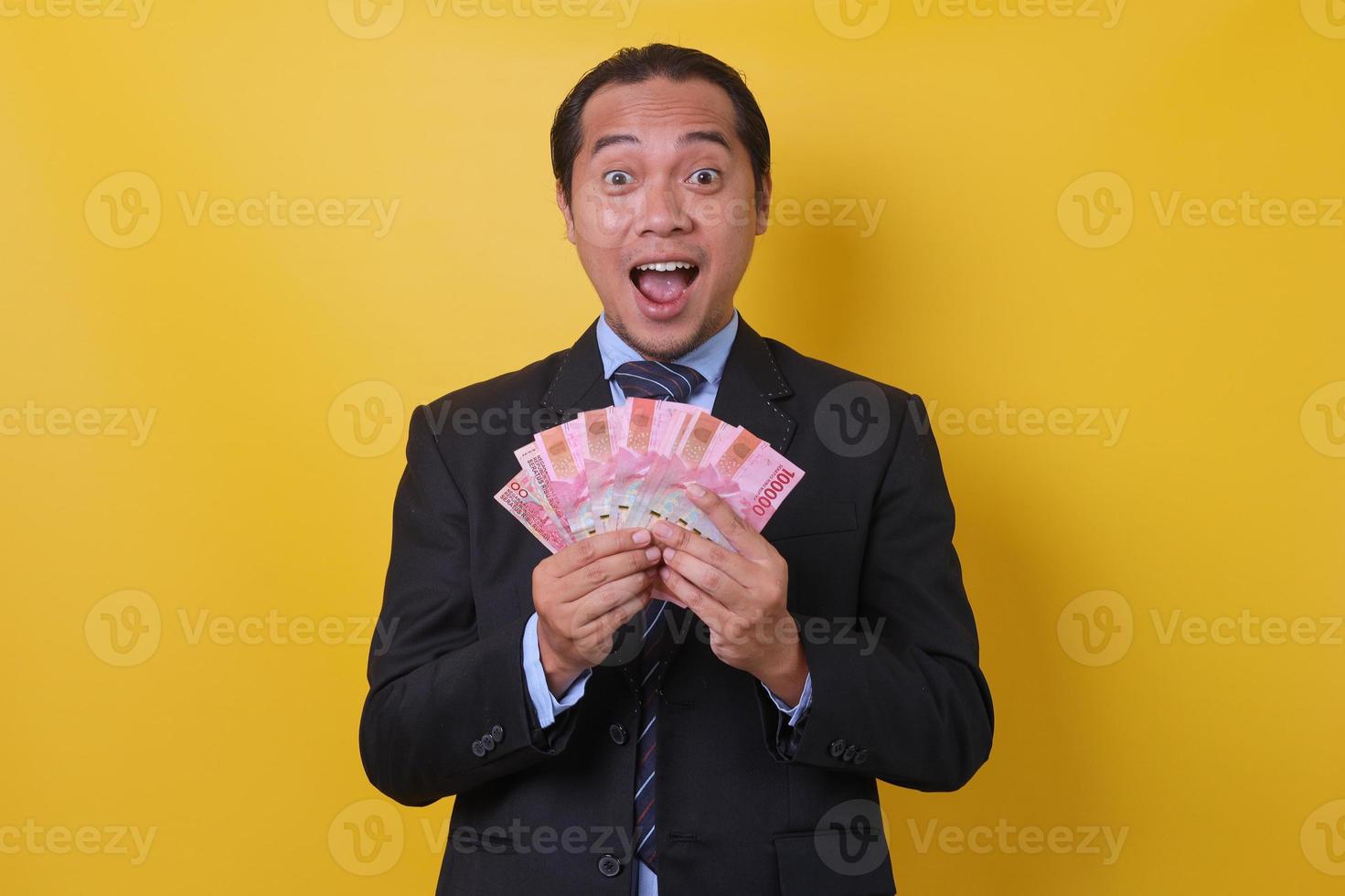 Portrait of excited Asian man in business suit standing against yellow background, showing one hundred thousand rupiah. Financial and savings concept. photo