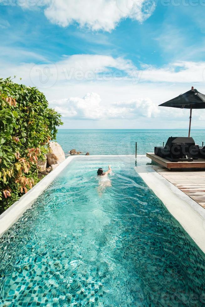 mujer feliz en traje de baño blanco nadando en la piscina infinita en un hotel de lujo frente al mar. mujer joven disfruta en un resort tropical. concepto de relajación, verano, viajes, vacaciones, vacaciones y fin de semana foto