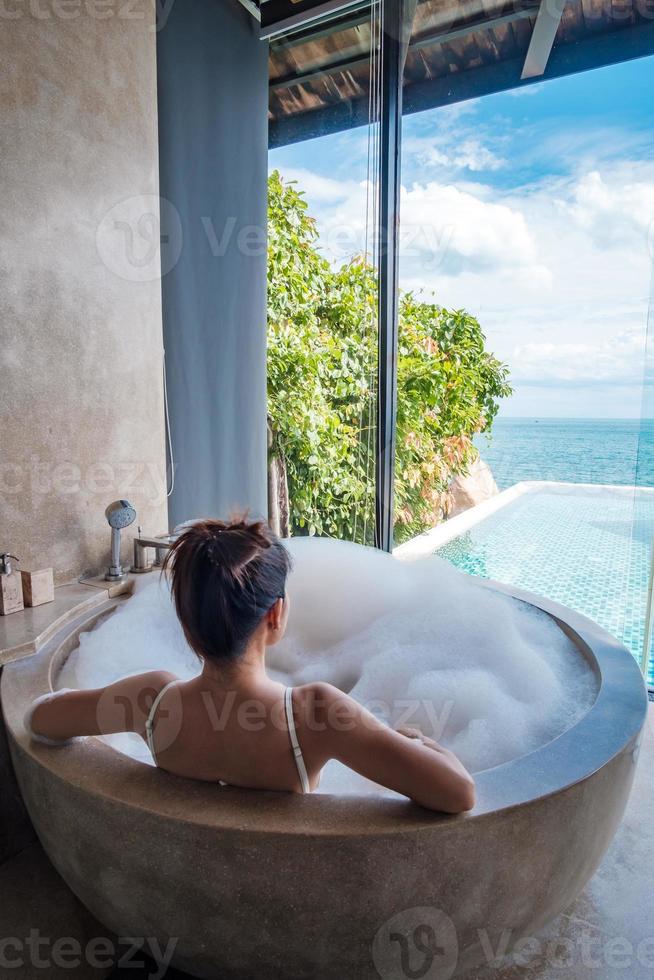 woman in white swimsuit relaxing in the bathtub with bubble foam against ocean background. Summer, relaxing and vacation concepts photo