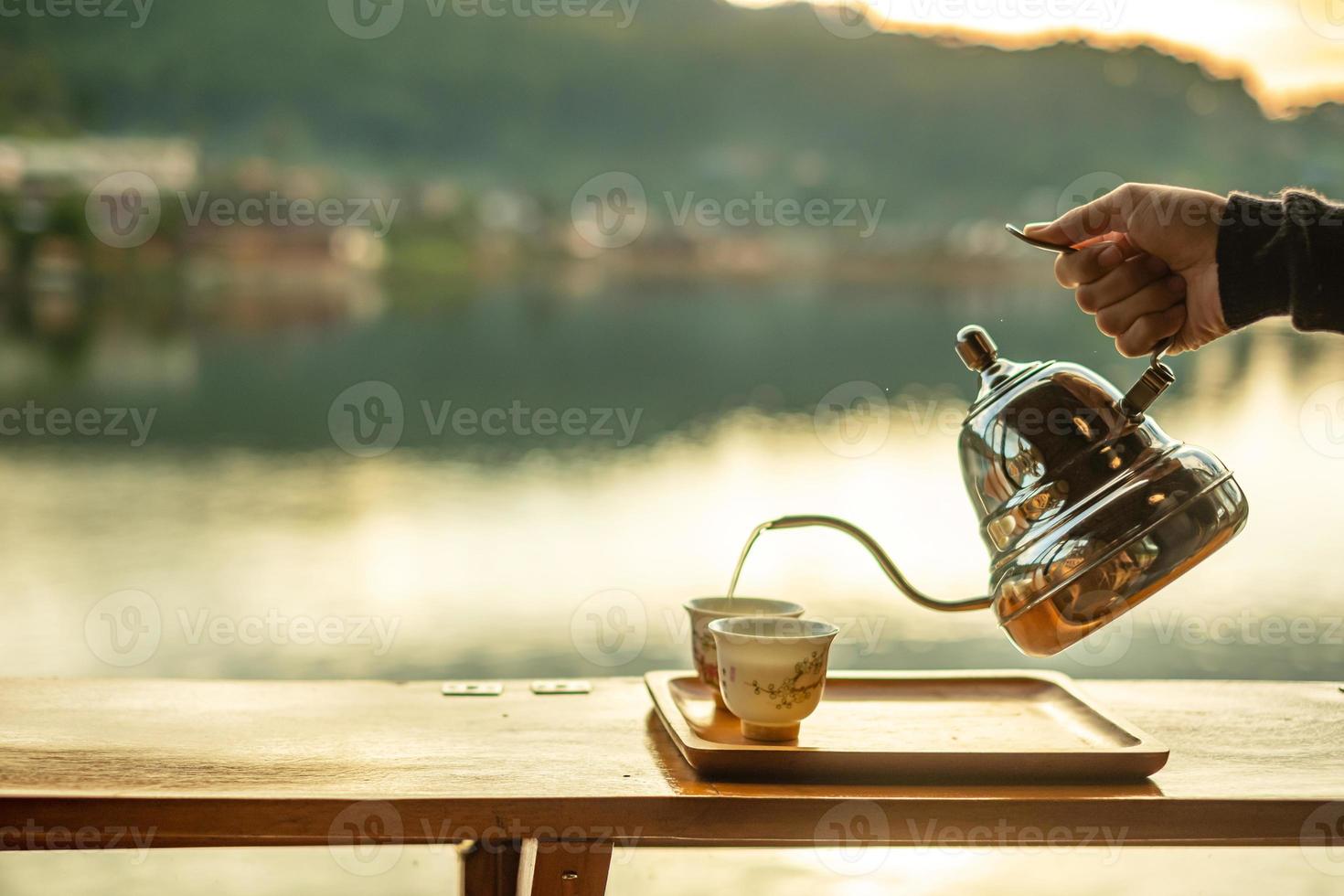 Hand holding vintage teapot and pouring hot tea to cup on wood table against lake view background at coffee shop in the morning sunrise, Ban Rak Thai village, Mae Hong Son province, Thailand photo