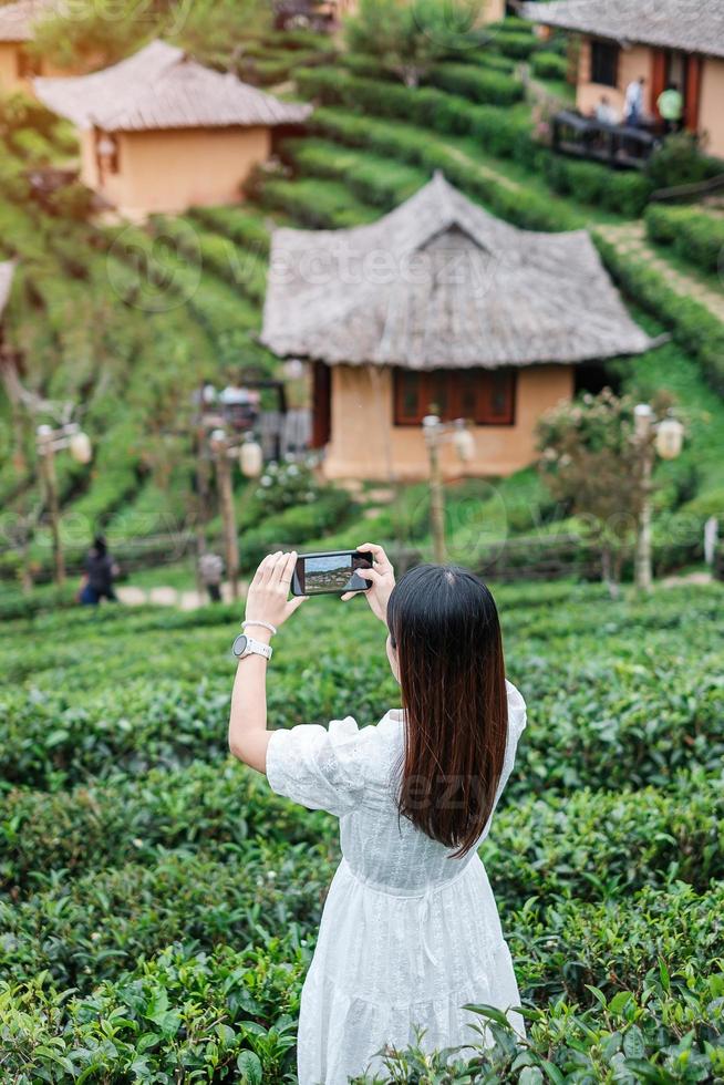 Happy tourist woman in white dress taking photo by mobile smartphone in beautiful Tea garden.Traveler visiting in Ban Rak Thai village, Mae Hong Son, Thailand. travel, vacation and holiday concept