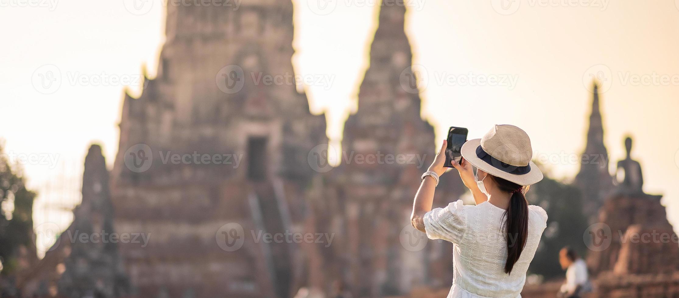 Happy tourist woman wearing surgical face mask and taking Photo by mobile smartphone, protection COVID-19 pandemic during visiting in Ayutthaya temple. new normal, safety travel and travel concept