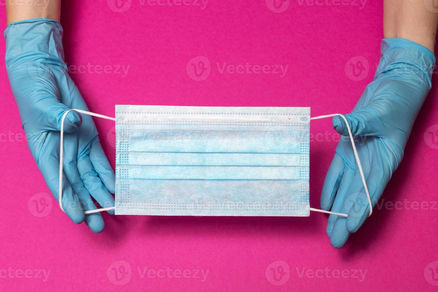 Hands in blue medical gloves is holding face mask on pink background. Protective respiratory medical mask against spreading virus. Close up, copy space, top view, flat lay. High quality photo