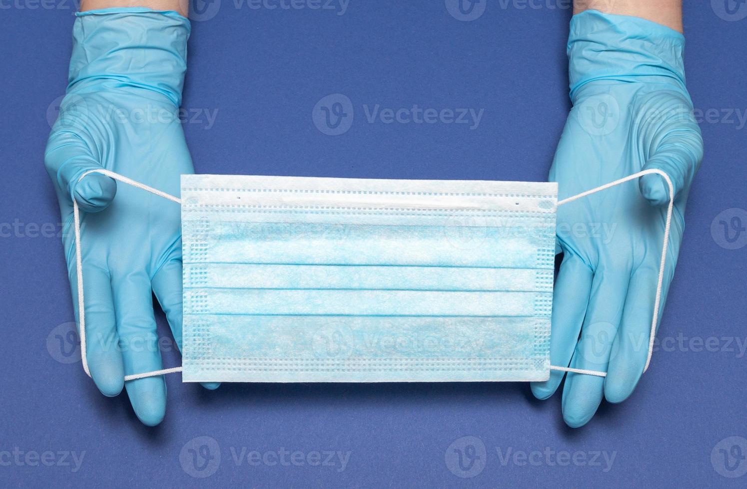 Hands in blue medical gloves is holding face mask on blue background. Protective respiratory medical mask against spreading virus. Close up, copy space, top view, flat lay. High quality photo
