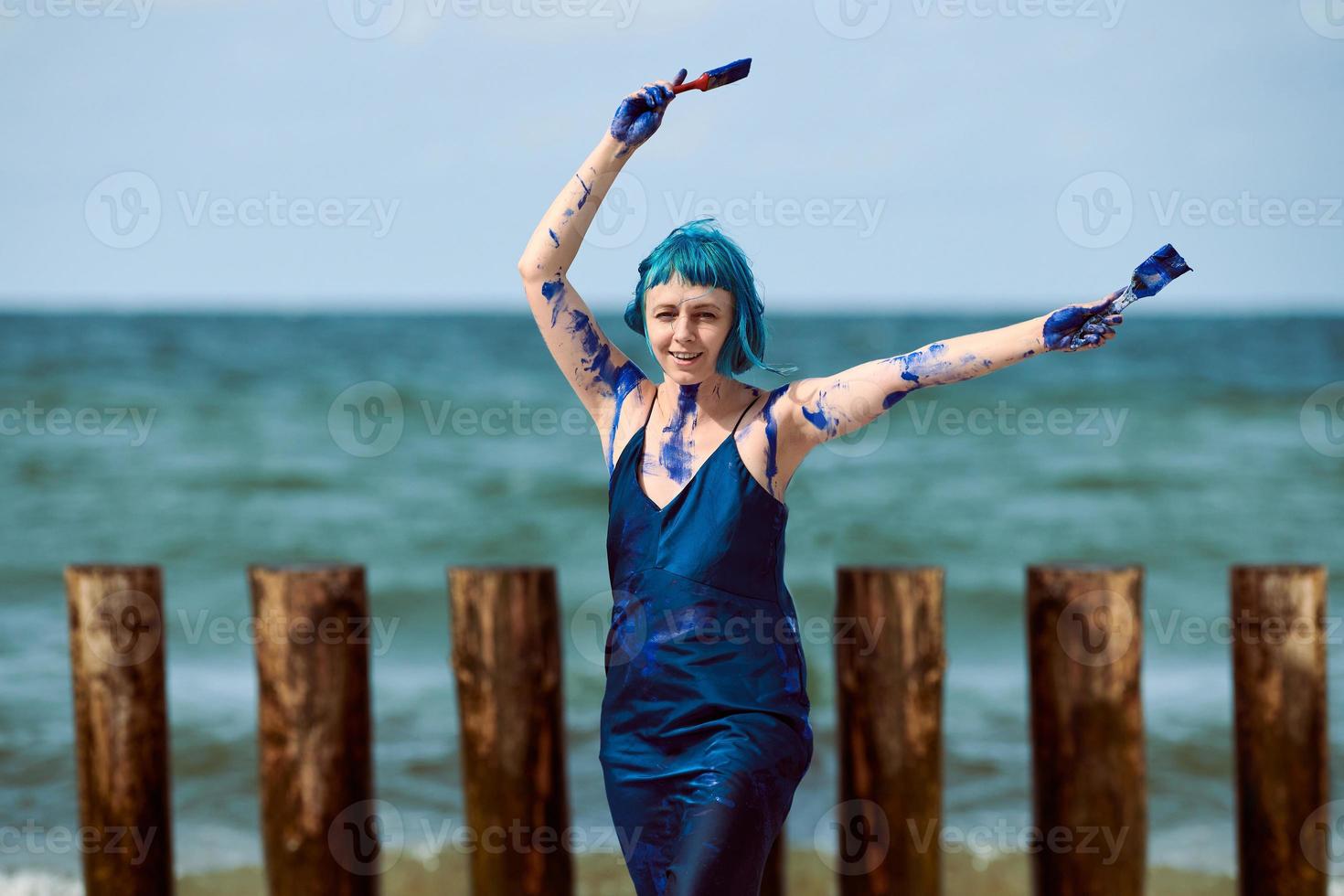 Artista de performance de mujer de cabello azul manchada con pinturas de gouache azul bailando en la playa foto